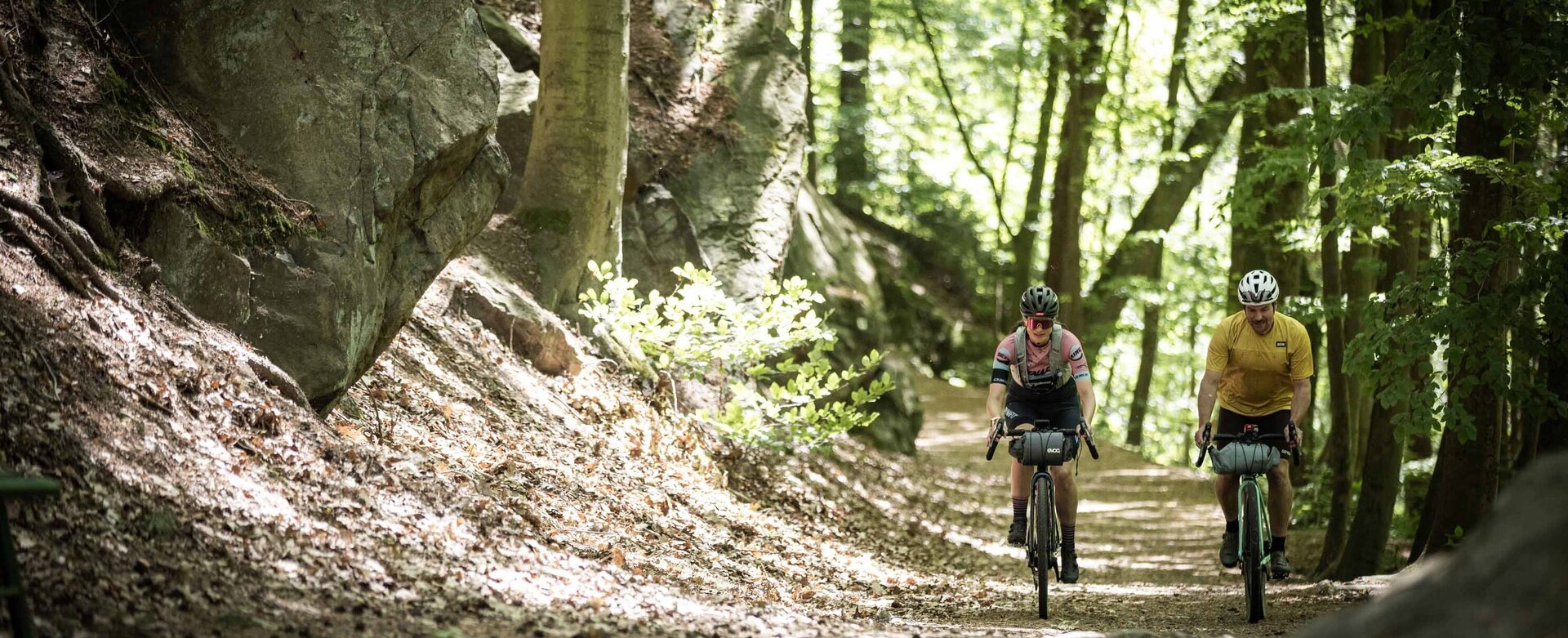 Zwei Gravelbiker fahren auf dem RockHead in der Nähe von Dürrröhrsdorf-Dittersbach durch einen Wald, links von ihnen sind Felsen zu sehen.