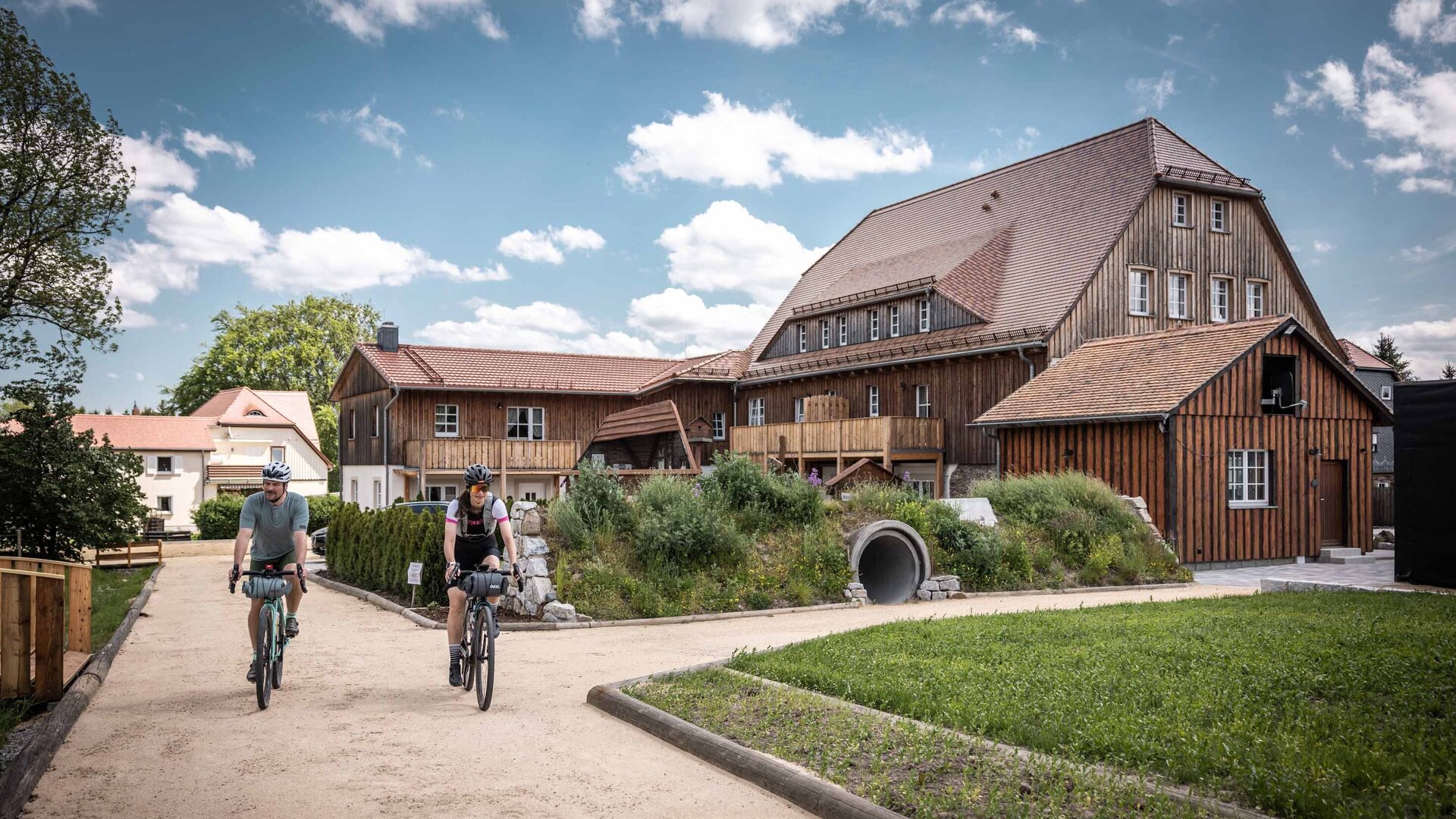 Zwei Gravelbiker fahren auf einem hellen Sandweg am Lerchenberghof in Eibau vorbei, einem markanten Holzgebäude mit Schindeldach und Balkonen. Die Radfahrer sind mit Bikepacking-Taschen ausgestattet und befinden sich auf der Rockhead-Route in der Oberlausitz. Im Hintergrund erstreckt sich ein blauer Himmel mit weißen Schönwetterwolken über der gepflegten Anlage des Ferienhofs.
