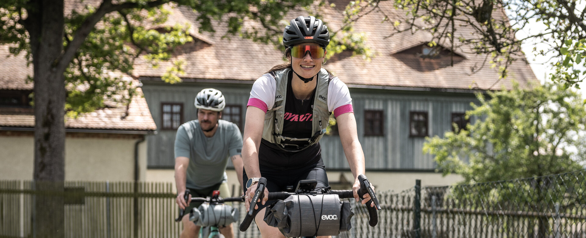Zwei Radsportler auf Gravelbikes befahren einen hellen Schotterweg am Faktorenhof in Eibau. Im Vordergrund lächelt eine Radfahrerin in sportlicher Bekleidung mit Helm und Sonnenbrille, während sie ihr Bike mit Lenkertasche sicher über den Pfad führt. Ein weiterer Fahrer folgt ihr in kurzem Abstand. Im Hintergrund ist die historische Architektur des Faktorenhofs mit seinen charakteristischen Hechtgauben und Holzfassaden unter sommergrünen Bäumen zu sehen.