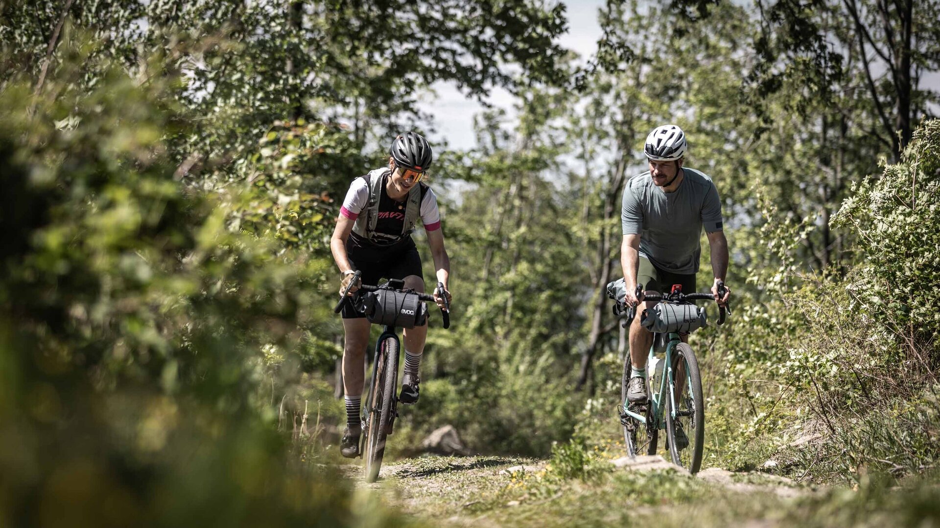 Zwei Gravelbike-Fahrer, eine Frau und ein Mann, fahren nebeneinander auf einem naturbelassenen Waldweg der Rockhead-Route in der Oberlausitz. Beide tragen Radsportbekleidung, Helme und haben Lenkertaschen für Bikepacking an ihren Rädern montiert. Die Szene ist von dichtem, grünem Laubwald umgeben, wobei der Fokus auf den konzentrierten Fahrern liegt, die eine leichte Steigung bewältigen.