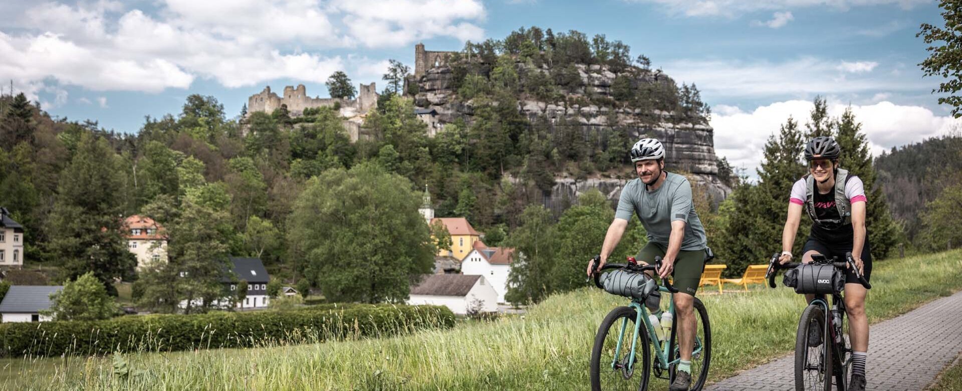 Zwei Gravelbiker fahren durch Oybin, im Hintergrund sieht man die Burgruine Oybin.