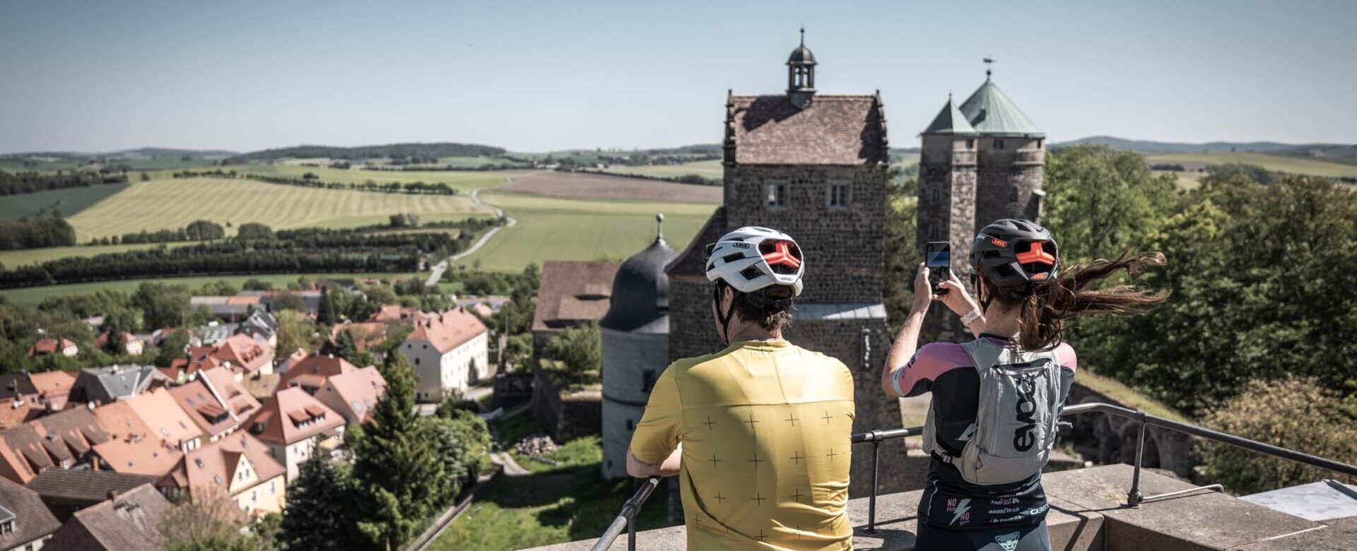 Zwei Gravelbiker stehen auf einem Turm der Burg Stolpen und machen Fotos von der Burganlage und der Aussicht über die Stadt.
